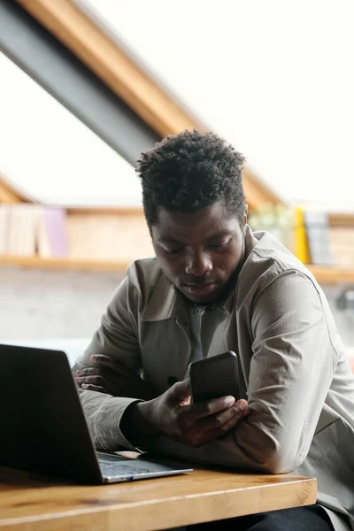 年轻的非洲裔美国商人坐在办公室的笔记本电脑前 一边用智能手机发短信 一边在屏幕上观看视频 — 图库照片