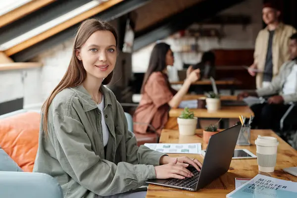 Young smiling female analyst or economist looking at camera while sitting by workplace in front of laptop and working with online financial data