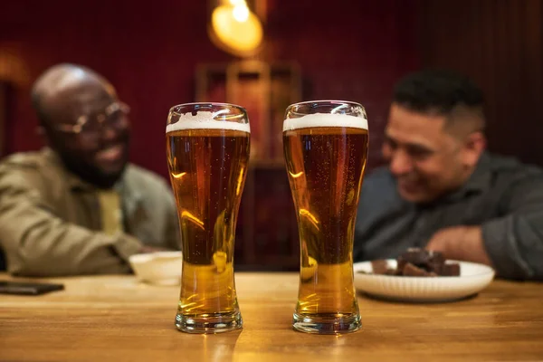 2つの異文化の仲間に対して木製のバーカウンターに立っている内部の小さな泡が付いている新鮮な涼しい泡立つビールの2つの大きいガラス — ストック写真