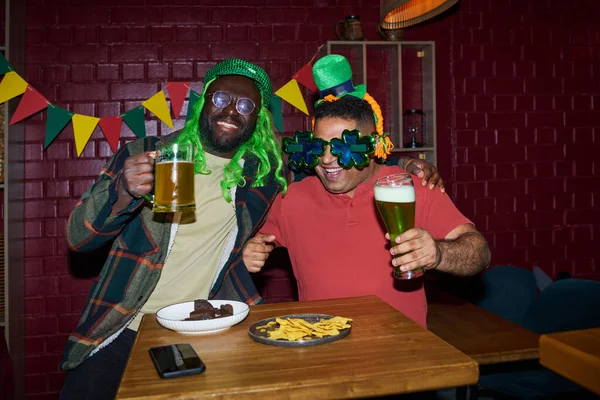 stock image Two cheerful intercultural buddies with glasses of beer toasting by table with snacks and looking at camera during celebration of St Patrick day