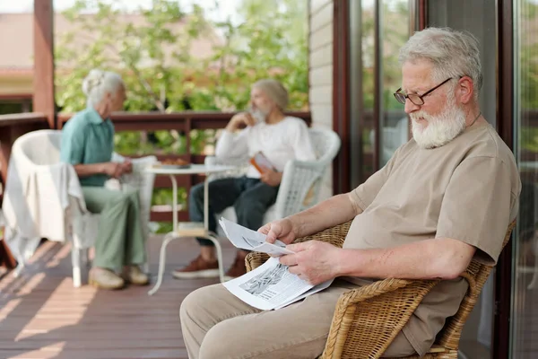 一个白发苍苍的大胡子老人坐在养老院的柳条椅上 一边看报纸 一边看最新消息 — 图库照片