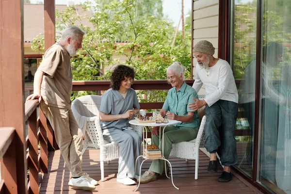 两个长胡子的老人坐在养老院的小桌旁 一边下棋 一边看着年老的女人和年轻的女人 — 图库照片