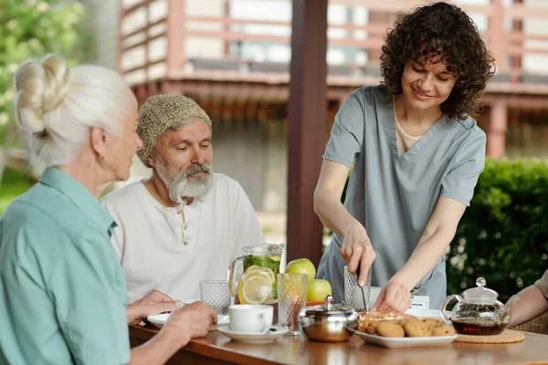 年轻漂亮的看护者为坐在桌旁的一群老年人切蛋糕或派 为他们提供早餐 并为他们端上甜点等茶 — 图库照片