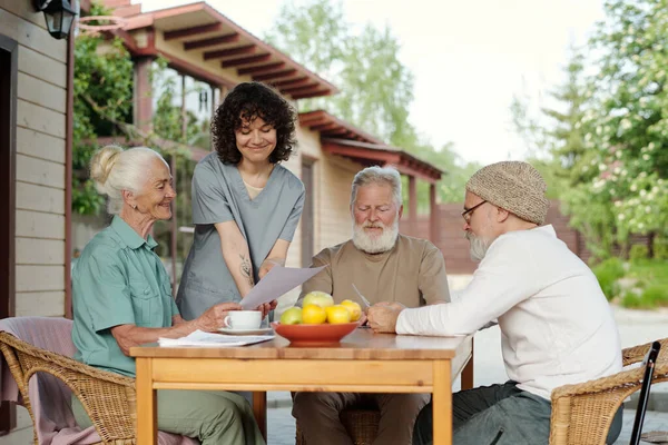 身穿制服 面带微笑的年轻护理者和老年妇女退休之家的病人 在看老年胡子男子旁边的纸质文件 — 图库照片
