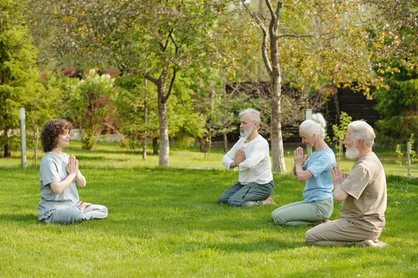 年轻女性瑜伽教师及退休老弱妇孺在青草地上练习冥想的侧视图 — 图库照片