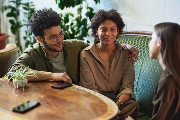Счастливая молодая межкультурная пара сидит на удобном диване за деревянным столом и смотрит на своего друга во время общения во время сбора в кафе