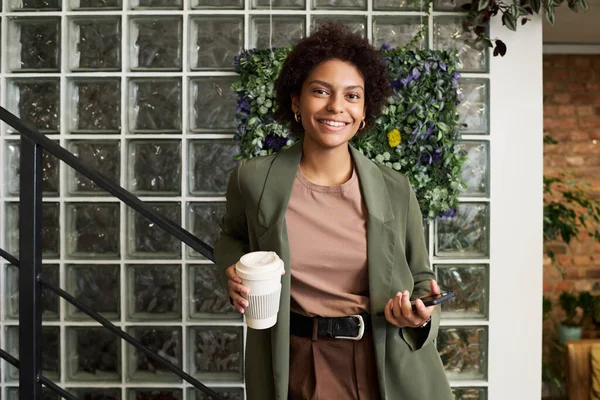 快乐的年轻女商人 拿着一杯咖啡 微笑着看着摄像机 手扶手靠着装饰着绿色植物的墙壁站着 — 图库照片