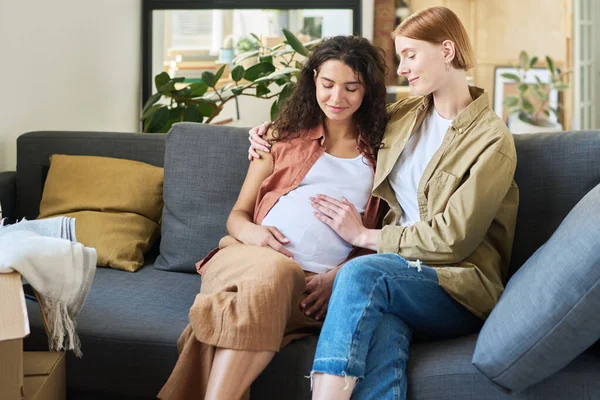 年轻的微笑的女人摸着怀孕的女朋友的肚子 拥抱着她 两个人坐在柔软舒适的沙发上 — 图库照片