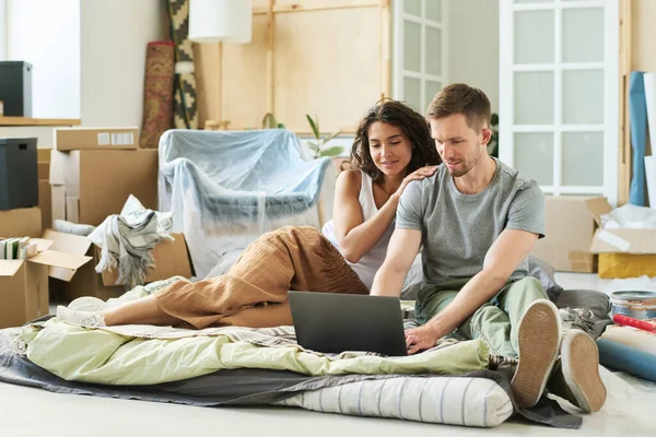 若い愛情のあるカップルは リビングルームの床に置かれたチェッカーに座って キーボードにタイピングしながらノートパソコンの画面を見ています — ストック写真