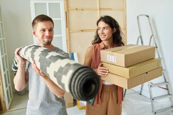 Happy young woman with stack of packed cardboard boxes looking at her husband with rolled carpet carpet while both moving along living room