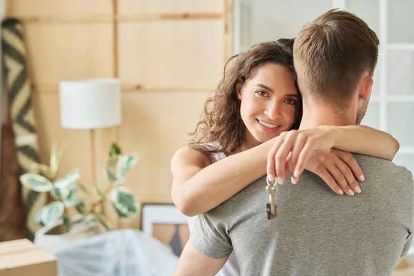 Happy young brunette woman with key from new apartment in hand embracing her husband and looking at camera after relocation
