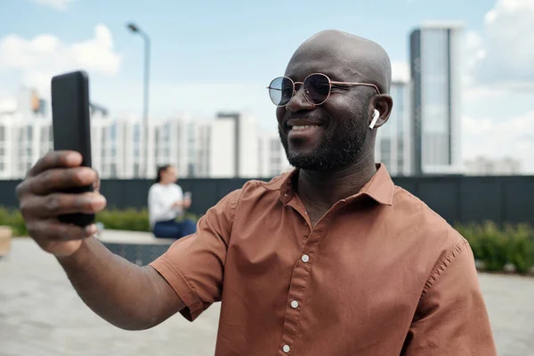 若い陽気なアフリカ系アメリカ人男性 イヤホンでスマートフォン画面を見て ビデオチャット屋外で誰かと通信しながら — ストック写真