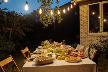 Ev yapımı yemek ve içeceklerle servis edilen masa arka bahçede kameranın önünde duruyor ve arka planda aydınlatma ve köy evi var.