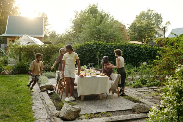 Intercultuiral family of five members preparing table in the garden for dinner while two siblings having fun and young man talking on phone