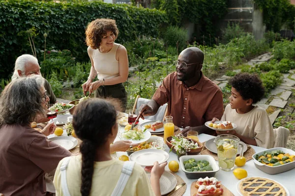 Happy young intercultural husband and wife, their two children and parents having outdoor family dinner in the garden or backyard