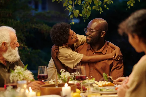 Adorable African American boy embracing his father and looking at him during outdoor family dinner by table served with homemade food