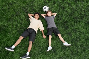 Yeşil çimlerde ya da futbol sahasında uzanan ve futbol oynadıktan sonra eğlenen hareketli kültürlü iki çocuğun yüksek açısı.