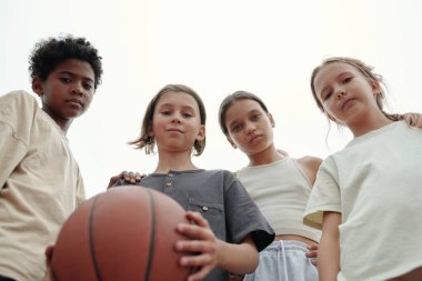Bir grup ciddi kültürler arası okul çocuğu kameraya bakarken bir tanesi açık hava maçında basketbol oynamak için topu tutuyor.