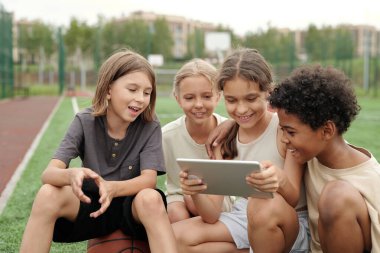 Kültürler arası mutlu sınıf arkadaşları online video izlerken gülümseyen sarışın kız tarafından tutulan tablet ekranına bakıyorlar.