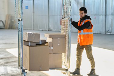 Depoda çalışan genç bir işçi içinde endüstriyel ekipmanlar için yedek parçaların bulunduğu paketlenmiş karton kutularla birlikte yük arabalarını itiyor.