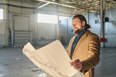 Geniş bir deponun ya da endüstriyel fabrikanın genç sakallı sahibi kameranın önünde duruyor ve bina planına bakıyor.