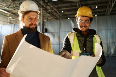 Genç gülümseyen mühendis inşaat planının detaylarını inşaat mühendisi veya ustabaşına açıklarken aynı zamanda iş toplantısının planına da işaret ediyor.