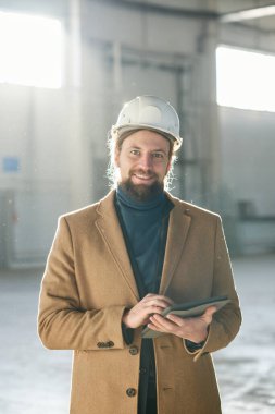 Güvenlik kaskı ve bej ceketli genç başarılı müteahhit ya da ustabaşı gülümseyerek kameraya bakıyor ve güneş ışığıyla aydınlanan iç mekâna karşı tablet kullanıyor.