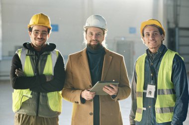 Koruyucu miğferli üç mutlu kültür mühendisi, ustabaşının etrafını saran güvenlik yelekleriyle kameraya gülümseyen iki adama bakıyor.