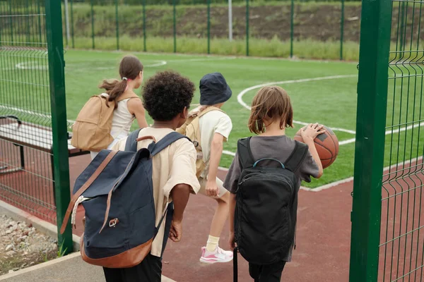 スポーツグラウンドやスタジアムに入る4人の子供のバックビュー そのうちの1人はバスケットボールをするためにボールをドリブル — ストック写真