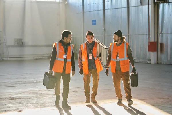 三个穿着工作服的跨文化工程师沿着仓库走着 其中两个拿着工具箱 看着带着台阶的同事 — 图库照片