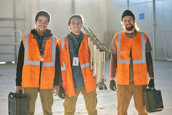 工場や倉庫の若い成功したエンジニアのグループは 日光に照らされたワークショップに立ちながら笑顔でカメラを見ています — ストック写真