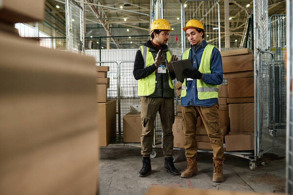 Young confident enginner in protective hardhat and reflective vest explaining something to colleague with tablet at working meeting