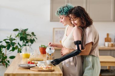 Genç ve mutlu esmer kadının, kahvaltı hazırlarken sevgilisini miyoelektrik eliyle kucaklamasının yan görüntüsü.