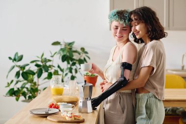 Sevecen genç bir kadın, mutfak masasının önünde elinde aperatiflerle dururken sevgilisini miyoelektrik koluyla kucaklıyor.