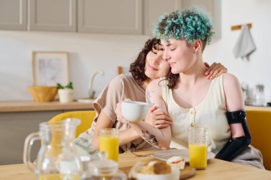 Genç esmer kadın miyoelektrik kollu kız arkadaşını kucaklıyor çay ya da kahve içerken ikisi de masada oturup kahvaltı yapıyor.