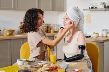 Genç esmer kadın, her ikisi de mutfak masasının yanında otururken gülümseyen kız arkadaşının tenine miyoelektrik bir elle bant yapıştırıyor.