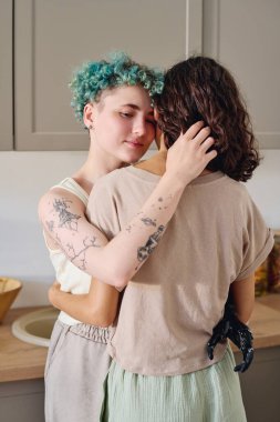 Engelli bir genç kadın, mutfak tezgahı ve dolapların önünde kameranın önünde dururken esmer kız arkadaşını kucaklıyor.