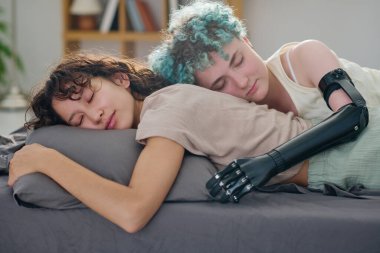 Myoelektrik elli, genç, sakin bir kadın. Uyurken kız arkadaşını kucaklıyor. İkisi de gri yatak örtüsüyle çift kişilik yatakta yatıyor.