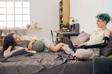 Özürlü genç bir kadın çift kişilik yatakta oturuyor ve hamile kız arkadaşının gri yatak örtüsünde yattığını izlerken dizüstü bilgisayarında klavye çalıyor.