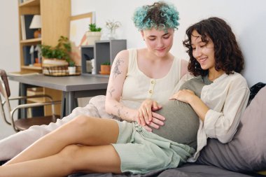 Miyoelektrik elli genç bir kadın gülümseyen hamile bir kız arkadaşın karnına dokunuyor. Bu arada ikisi de çift kişilik yatakta oturup birlikte vakit geçiriyorlar.