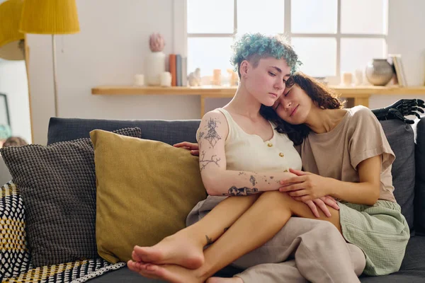 stock image Young serene lesbian couple in casualwear enjoying rest on comfortable couch with cushions against window and interior of living room