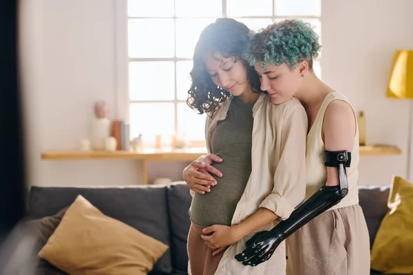 Young female amputee embracing her pregnant girlfriend and touching her belly while both standing in front of camera against couch