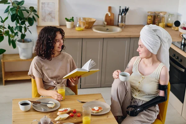 Genç sırıtan esmer kadın, açık bir kitapla mutfak masasında oturmuş kahvaltıda sohbet ederken engelli kız arkadaşına bakıyor.