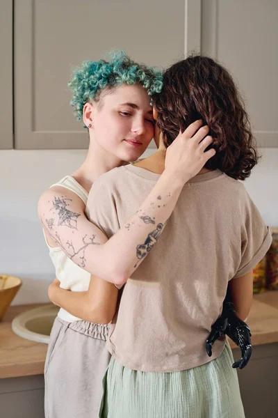 Engelli bir genç kadın, mutfak tezgahı ve dolapların önünde kameranın önünde dururken esmer kız arkadaşını kucaklıyor.