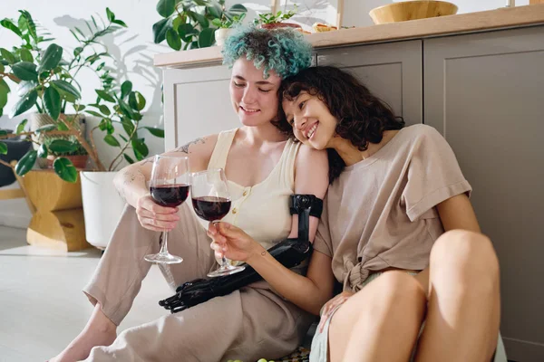Young cheerful amputee woman with glass of red wine toasting with her pretty brunette girlfriend while both sitting by kitchen counter