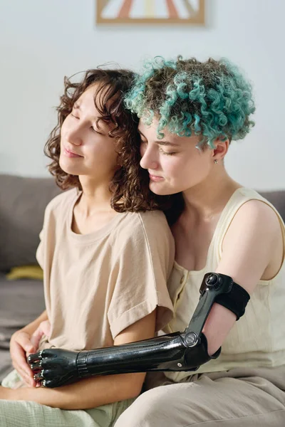Young female amputee with short blue curly hair embracing her pretty brunette girlfriend while sitting close to her in front of camera