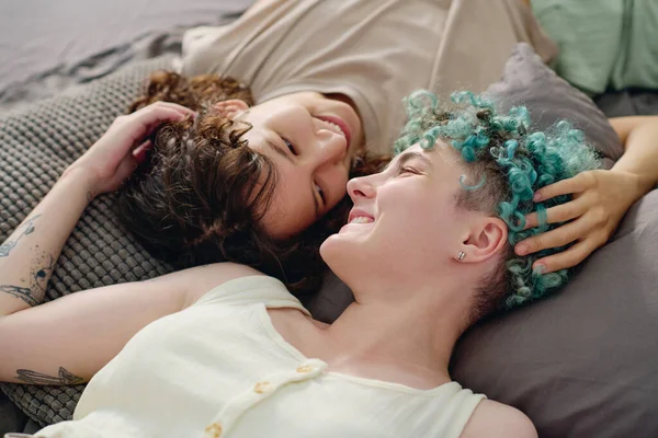 Evdeki gri yatak örtüsüyle yatakta dinlenirken dişlek tebessümlerle birbirlerine bakan kültürler arası neşeli kızlar.