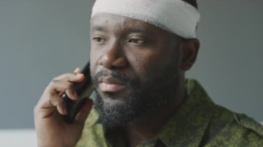 Kafasından yaralanmış sakallı Afrikalı Amerikalı yüzbaşının yakın plan portresi. Hastane odasında oturmuş telefonla konuşuyor.