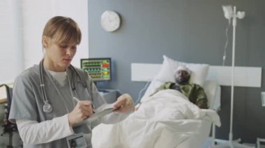 Askeri hastanede doktor olarak çalışan genç beyaz kadın baygın bir hastaya karşı duruyor ve tıbbi kartıyla bir şeyler yazıyor.
