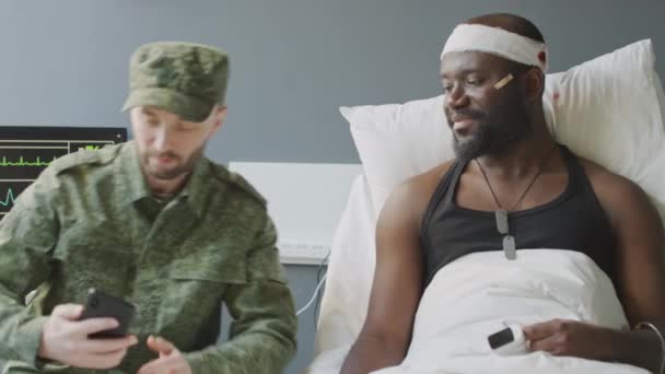 2人の民族的に多様な兵士が軍病院の部屋でスマートフォンでセルフィー写真を撮って戦闘で生き残った — ストック動画